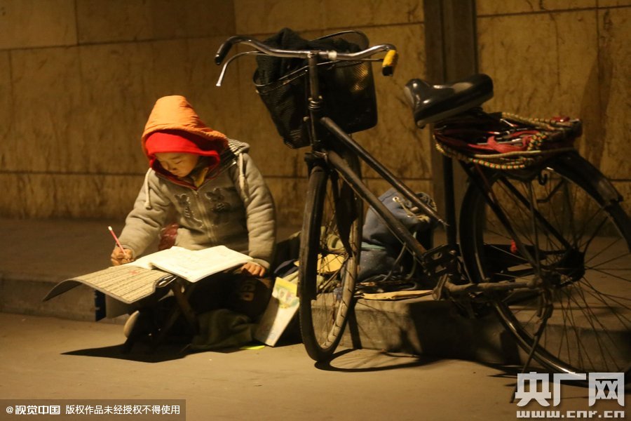 鄭州中原路京廣鐵路大橋下涵洞的馬路牙子邊，一位戴著紅色辣椒帽的小男孩趴在一個小馬扎上，上面鋪著一張車墊，在微弱的燈光下寫作業(yè)
