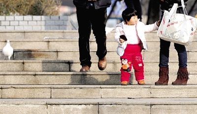 昨天中午，一女孩拉著家長的手在紫荊山公園曬太陽，氣溫升高，孩子的棉衣解開了扣子。